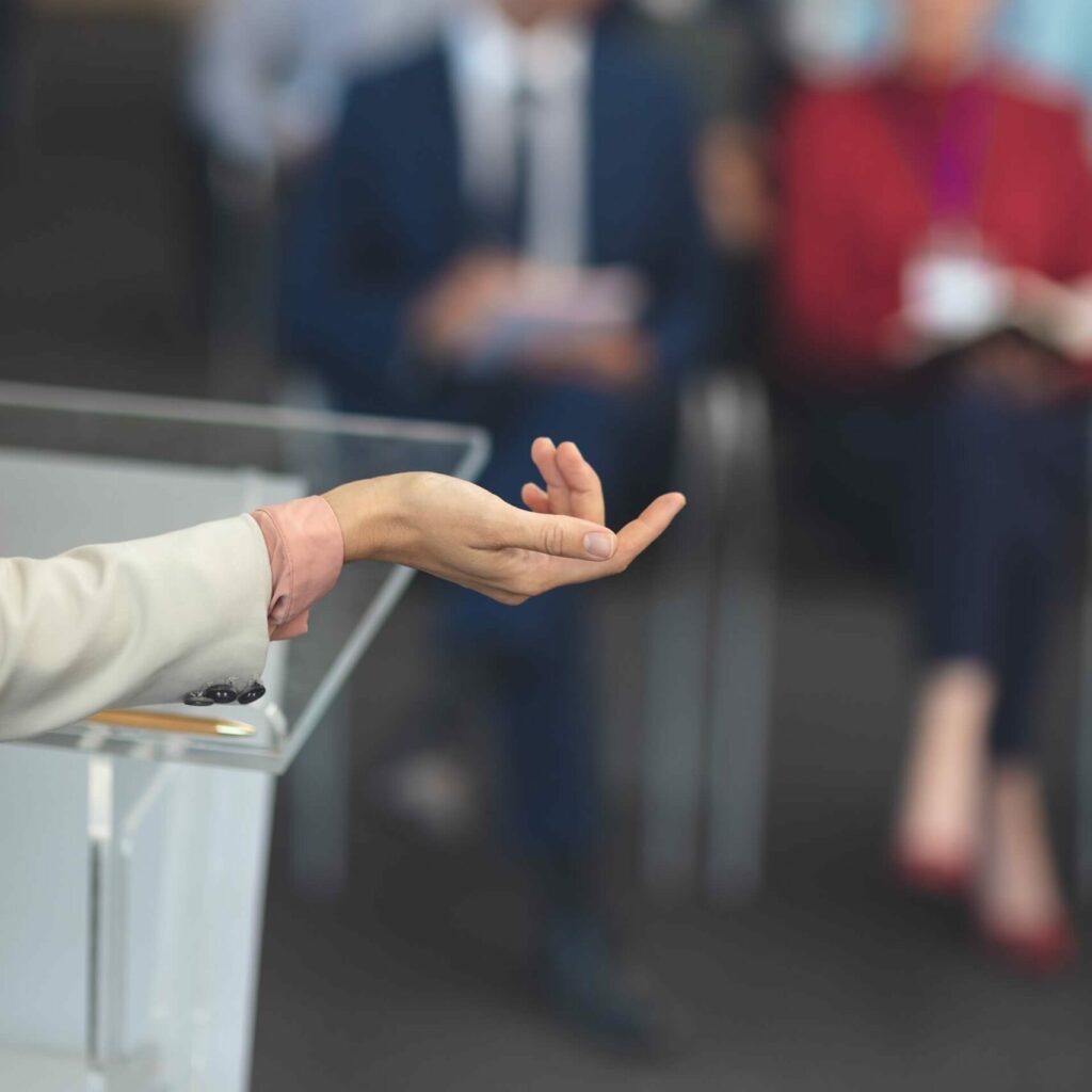 A close up photo of a female speaker's hand with a blurry background of an audience